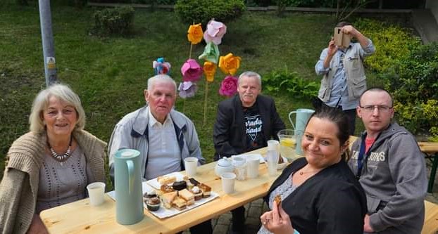 Na zdjęciu widać Uczestników i ich rodzinę siedzących przy stole i uśmiechających się w tle trawa i ogród.
