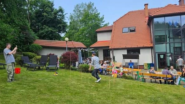 N azdjęciu widać trawnik, na którym dwuch Uczestników przygotowuje tor przeszkód. Na drugim planie stoły z poczęstunkiem w tle budynek Ośrodka.