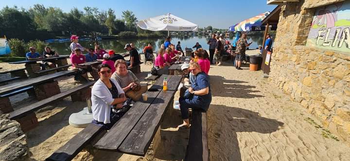 Na zdjęciu widać trzech Uczestników siedzących na dworze przy drewnianej ławie, w tle plaża i jezioro.