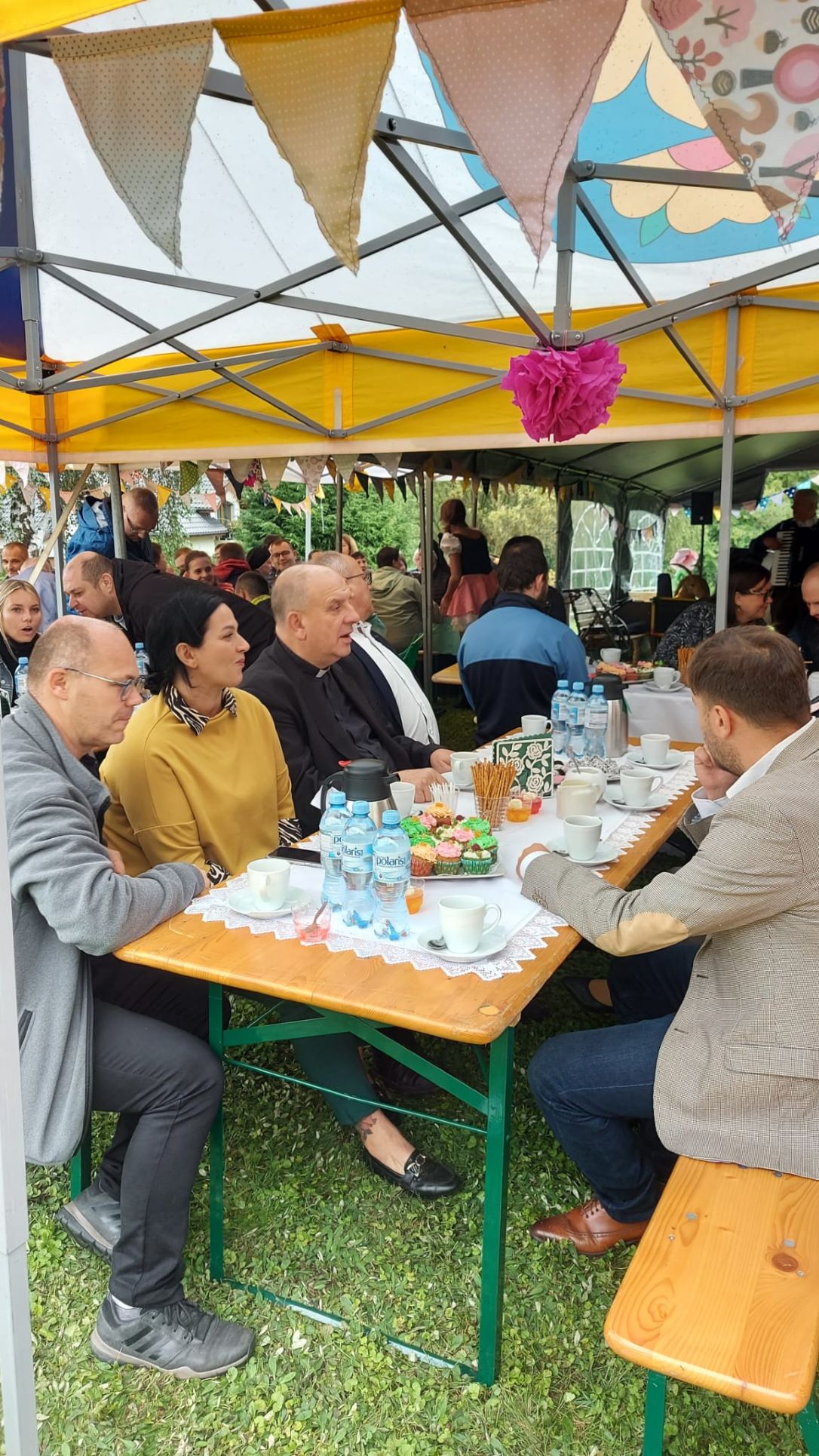 Grupa osób siedząca przy stole, na którym stoi kawa, woda, ciasta, w tle inne stoły i ławki oraz siedzące przy nich osoby.