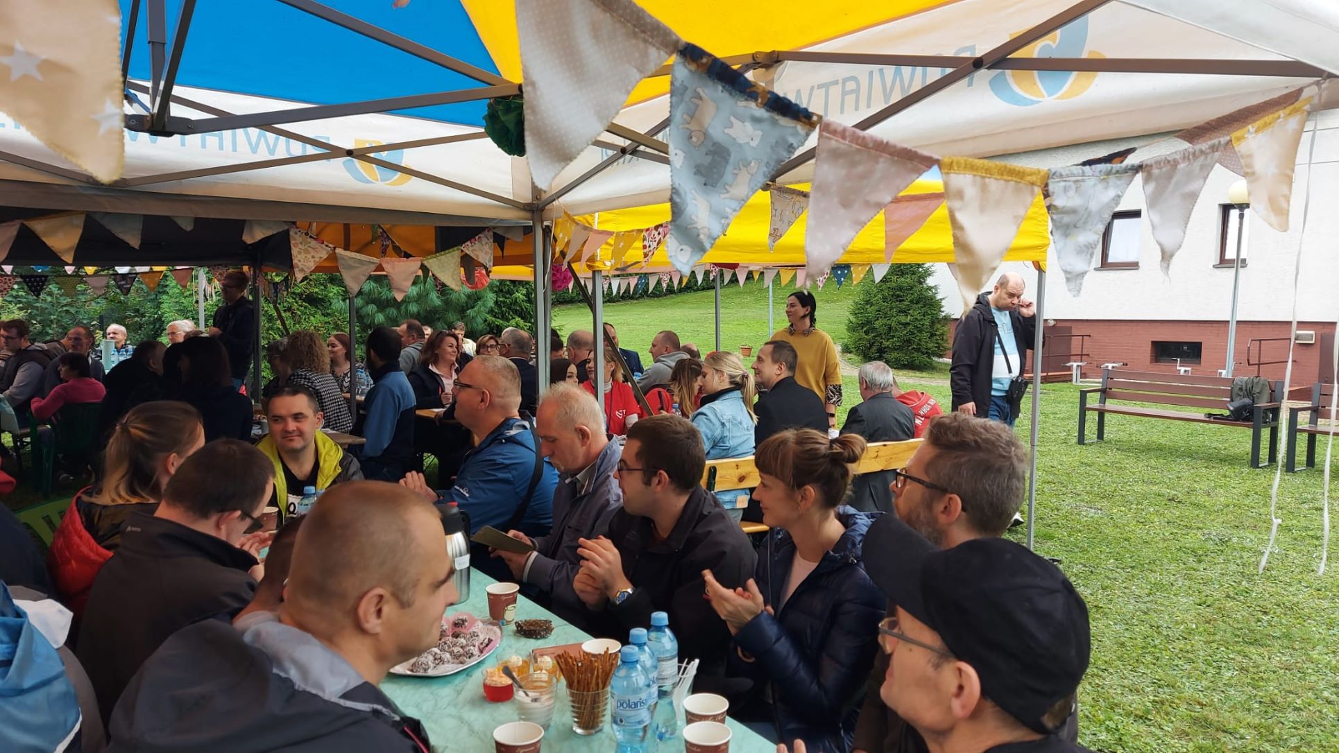 Grupa osób klaszcząca w dłonie siedząca przy stole, na którym stoi kawa, woda, ciasta w tle podwórk, trawa, fragment budynku.