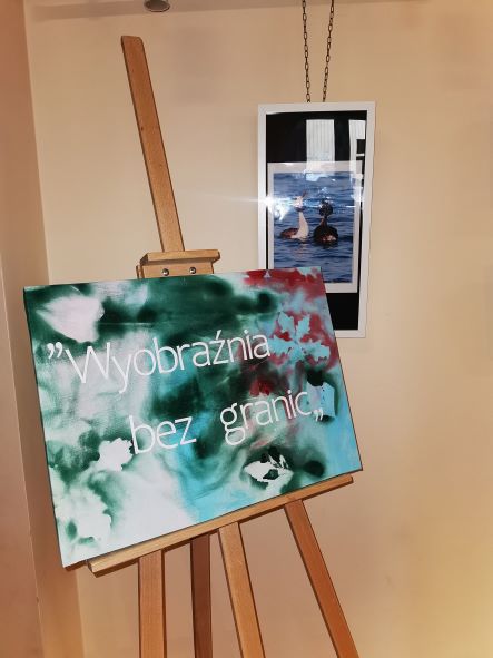 Na pierwszym planie stoi sztaluga z plakatem, na którym widać tytuł wystawy "Wyobraźnia bez granic", w tle zdjęcie przedstawiuające dwa perkozy.