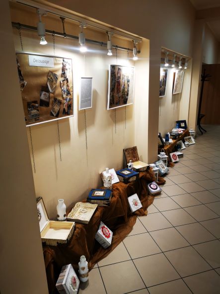 N zdjęciu widać wystawę prac uczestników, na ścianie wiszą antyramy ze zdjęciami, na podwyższeniu stoją pudełka i tacki wykonane techniką decoupage.