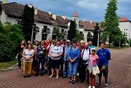 Na zdjęciu widać grupę kilkunastu osób pozującą do zdjęcia na tle dużego białego budynku z czerwonym spadzistym dachem.