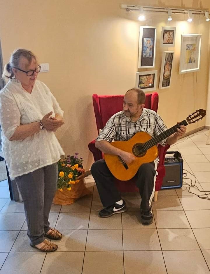 Na zdjęciu widać Uczestnika siedzącego na czerwonym fotelu grającego na gitarze i stojącą obok Uczestniczkę klaszczącą i śpiewającą.