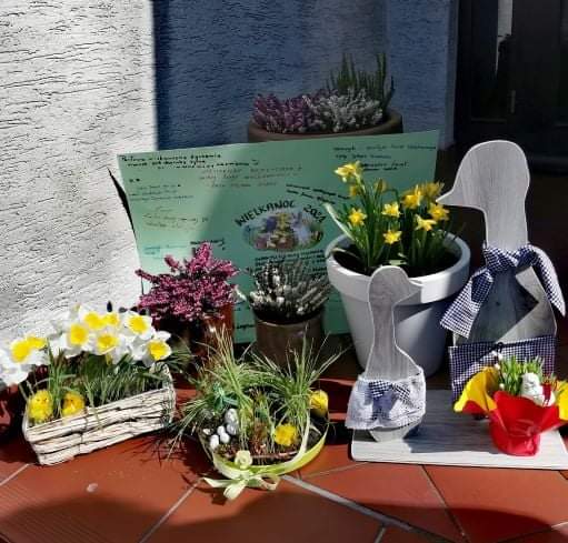 Na zdjęciu widać stroiki z żonkilami, trawą i kurczaczkami oraz drewniane ozdoby w kształcie kaczek. W tle karton, na którym uczestnicy pisali życzenia świąteczne.